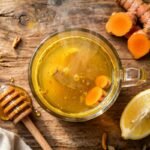 Glass mug of golden turmeric tea with fresh turmeric slices, lemon wedge and honey dipper