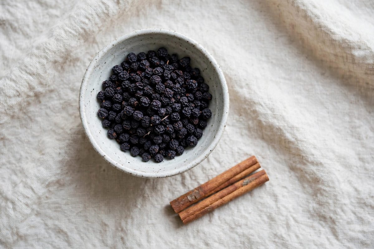 Top-down view of a ceramic bowl of dried dark purple-black elderberries on a linen cloth with cinnamon sticks
