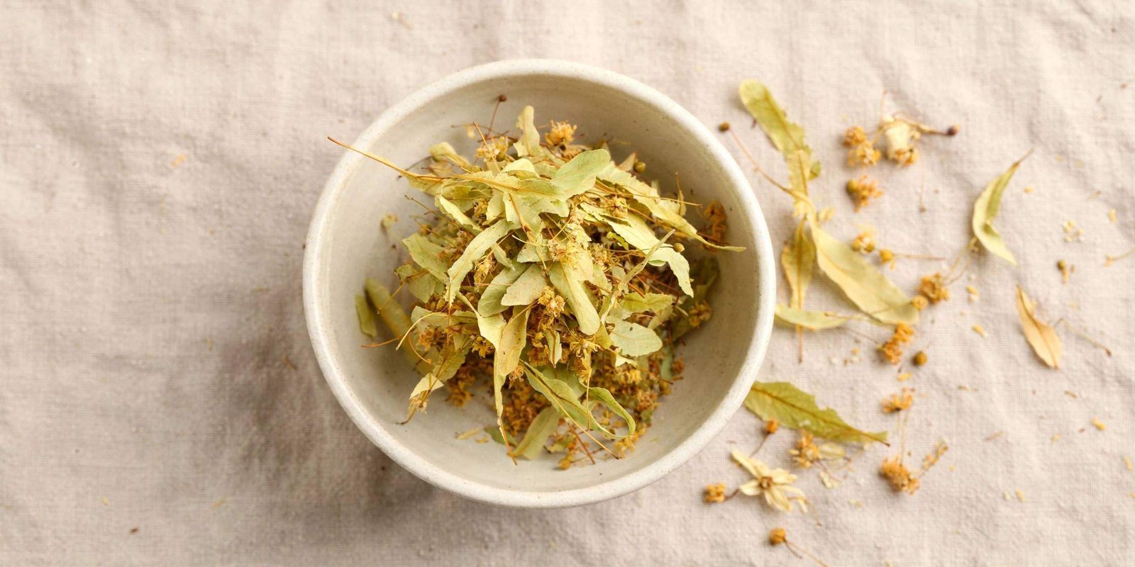 Top-down view of a ceramic bowl of dried linden flowers with their pale papery bracts on a linen cloth