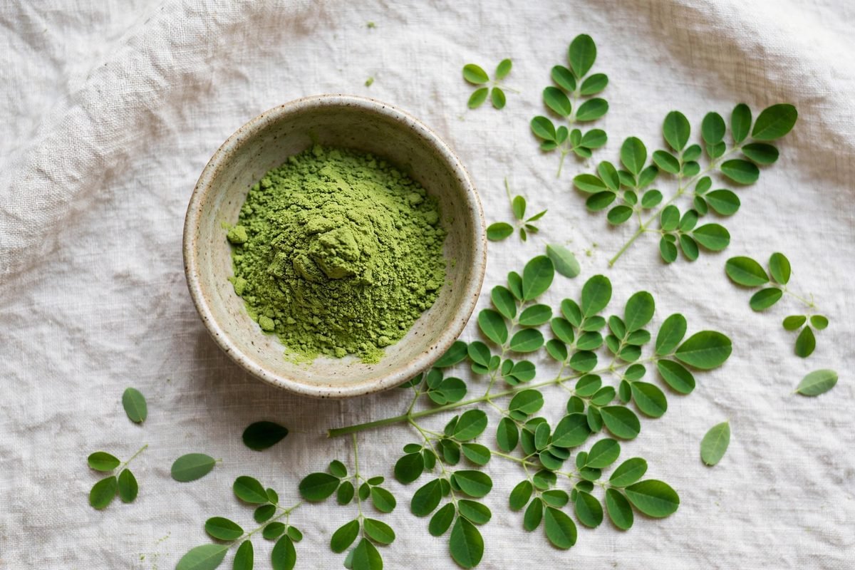 Top-down view of a ceramic bowl of green moringa powder beside fresh moringa leaves on a linen cloth